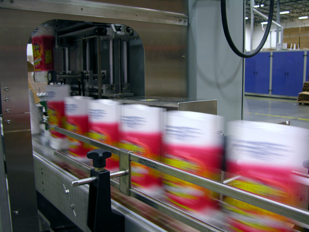 A processing plant showing automated machine attaching shrink sleeve labels to a packaging container.